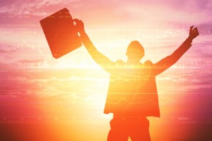 A forex trader raising his arms in joy with a beautiful sunrise behind him.